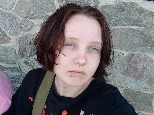 OliviaFlex, a young woman with pale skin and short, messy brown hair, stands against a stone wall. She has blue eyes, a small nose with a silver nose ring, and a lower lip piercing. She wears a black shirt with a red pattern. An olive-green strap crosses her chest. The stone wall in the background is rough and gray, with varying textures. Her expression is neutral, and she looks directly at the camera. The image is a close-up, focusing on her face and upper body.