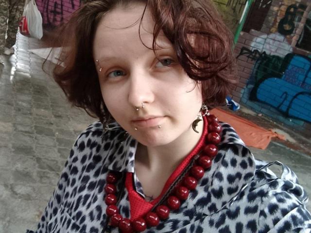OliviaFlex, a pale-skinned woman with wavy, brown hair, stares into the camera with a neutral expression. She has multiple facial piercings: a small stud on her right upper lip and a barbell on her lower lip. She wears a nose ring and has small earrings. Her outfit features a leopard-print shirt with a red beaded necklace. The image is a close-up, focusing on her upper body and face. The background is blurred, showing a graffiti-covered wall. The texture of the street and the graffiti adds urban detail.