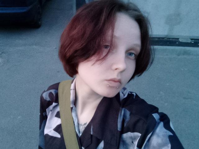 OliviaFlex has short, dark red hair and light skin. She has a small silver nose piercing on her left nostril and slightly parted lips. She wears a black and white patterned shirt with a beige strap across her shoulder. Her blue eyes look off to the side. The image has a cool, bluish tint. The texture of the shirt is slightly shiny. The background is a concrete pavement and a dark blue car.