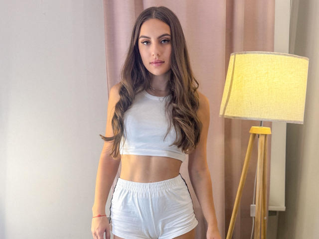 MaddymidNight with long brown hair, white sports bra, and white shorts, standing in front of a yellow lamp, showing a toned midsection and a neutral expression.
