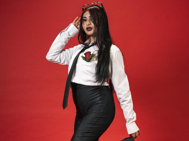 EmmaaRodriguez with long black hair, wearing a white shirt, black tie, red rose, black skirt, and sunglasses on head, holding a phone, standing against a red background.
