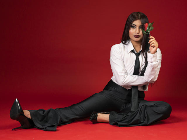 EmmaaRodriguez with dark makeup, white shirt, black tie, black pants, and high heels, sitting on red, holding a rose, showing her ass, and looking at the camera.