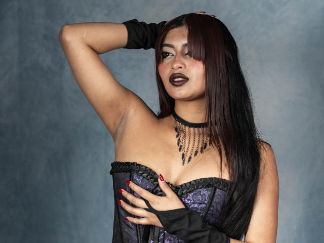 EmmaaRodriguez with dark makeup, black gloves, purple lace corset, and chain necklace, hand on her tits, red nails, and brown hair, looking seductive.