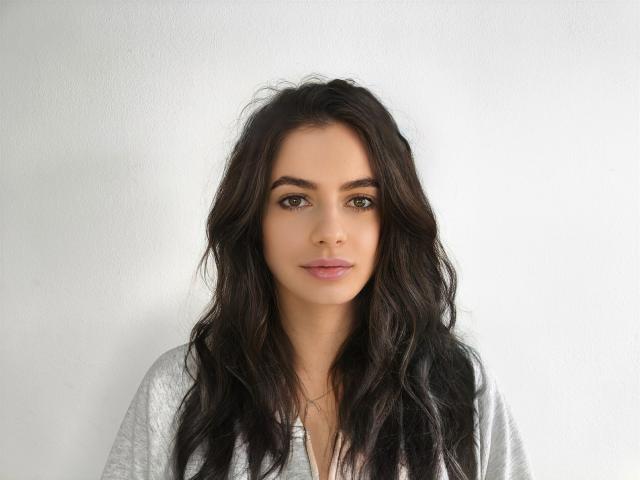 CarliHot has long, wavy dark brown hair, green eyes, and is wearing a light gray shirt. She's looking straight at the camera with a subtle, calm expression.