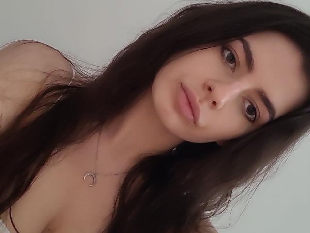 CarliHot with long, dark brown hair, brown eyes, and pink lips, wearing a thin necklace with a small crescent moon pendant, has a slight smile and is looking at camera.