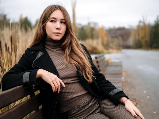 AmmaLoyXX with long brown hair, wearing a black coat, brown turtleneck, and gray patterned pants, sitting on a wooden bench by a river, looking at the camera.