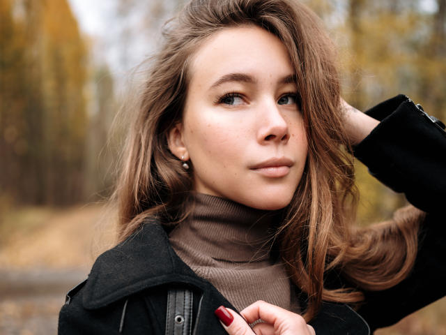 AmmaLoyXX has brown hair, light skin, and is wearing a black jacket over a brown turtleneck. She's looking at the camera with a slight smile, hand in hair.