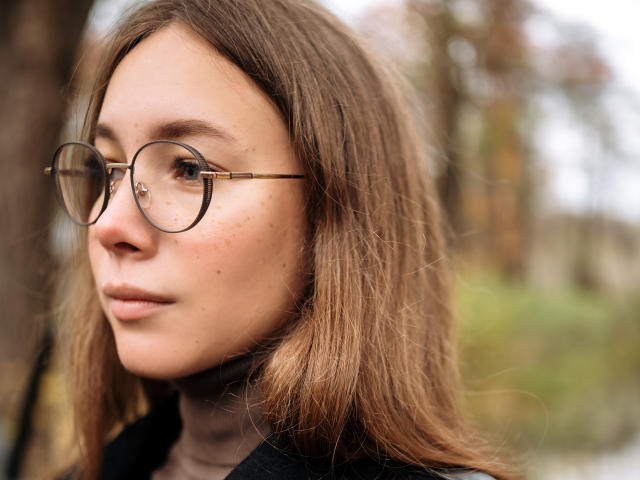 AmmaLoyXX has round glasses, brown wavy hair, and freckles on her face, wearing a black turtleneck, with a serious expression, in a soft, natural light.