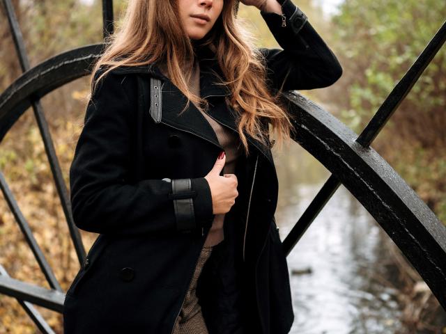 AmmaLoyXX with long brown hair, wearing a black coat, standing by a black metal structure, hand in coat, visible tits, wet ground, autumn forest background.
