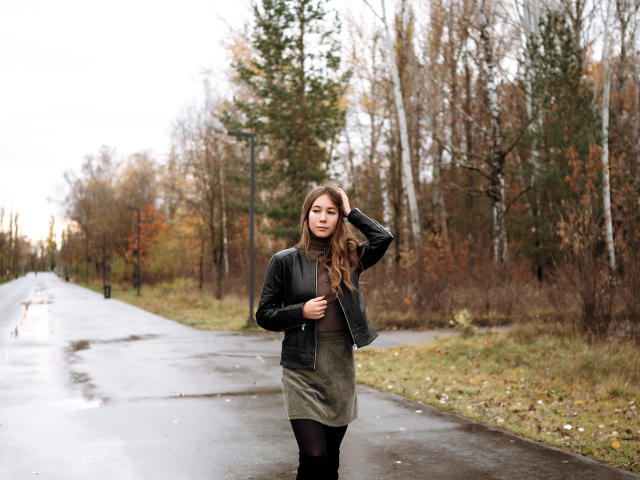 AmmaLoyXX has long brown hair, wearing a black leather jacket, brown turtleneck, green skirt, and black tights, standing on a wet, empty road with trees in the background.
