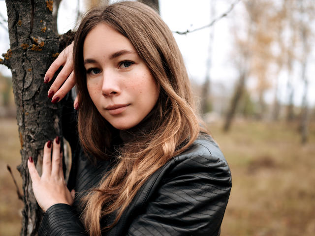 AmmaLoyXX with long brown hair, freckles, and red nails, leaning on a tree, wearing a black quilted jacket, looking at the camera with a subtle smile.