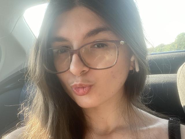 MarrieS has brown hair, wears glasses, and pouts her pink lips. Her ears have small silver earrings. Sunlight highlights her clear skin. She's sitting in a car.