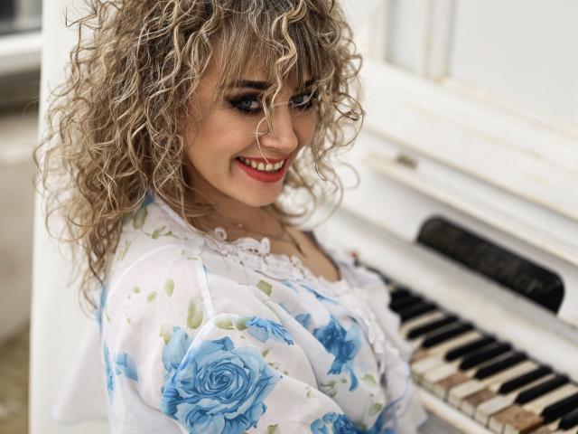 CandyLadyZ has curly blonde hair, wearing a white floral dress with blue roses. She's smiling with red lipstick, looking over her shoulder. She's standing next to a white, slightly worn piano with black and white keys. The image uses natural light, highlighting her fair skin and blue eyes. The focus is on CandyLadyZ's face and upper body. The texture of her hair is loose and curly, contrasting with the smooth fabric of her dress. The piano is slightly out of focus.