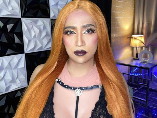 JuicyHugeCockTS with long orange hair, dark lipstick, and heavy makeup, wearing a black lace top with a pink necktie, and a beaded necklace, has a serious expression.