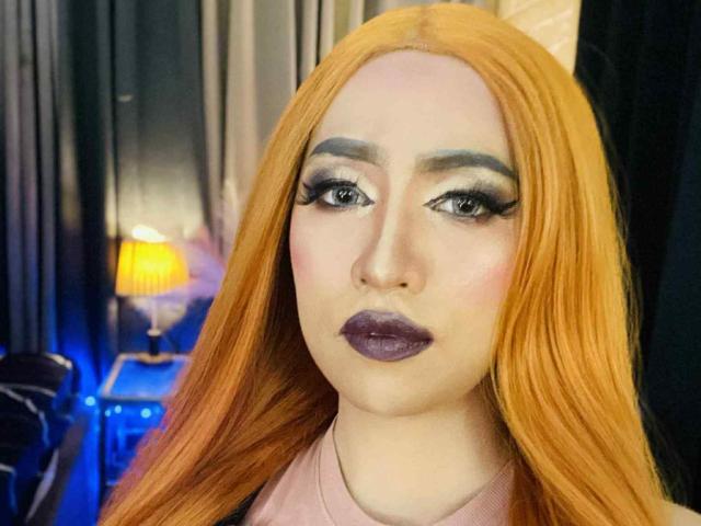 JuicyHugeCockTS with long orange hair, thick black eyebrows, heavy eyeliner, and purple lipstick, looking directly at the camera, wearing a pink shirt.