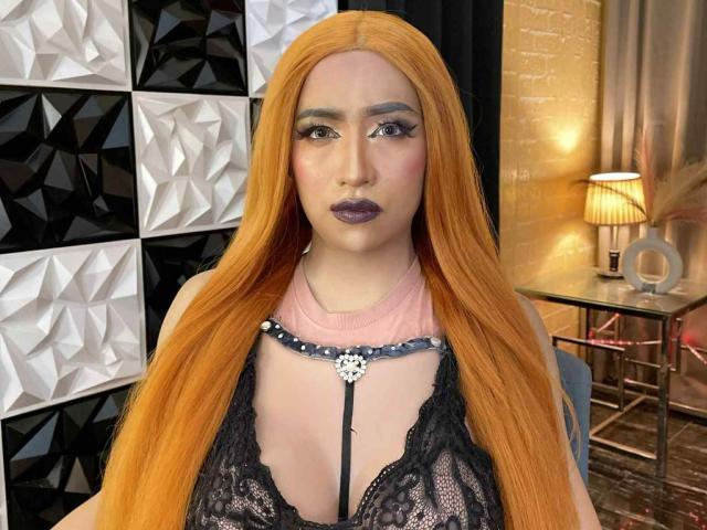 JuicyHugeCockTS with long orange hair, dark purple lipstick, black lace top, visible cleavage, and a pink collar with a silver ring, standing in a well-lit room.