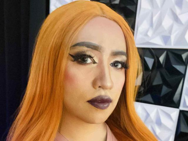 JuicyHugeCockTS has long orange hair, dark makeup, thick eyeliner, and purple lipstick. She's staring at the camera with a serious expression. Her eyes are wide and brown.