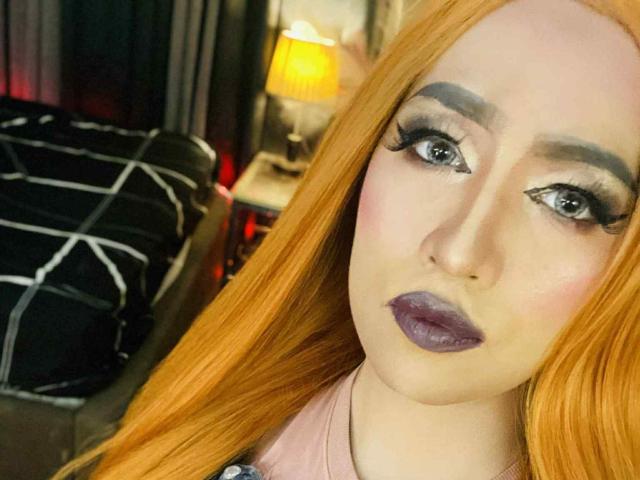 JuicyHugeCockTS with long red hair, purple lipstick, and heavy eyeliner, looking at the camera with a neutral expression, wearing a pink shirt.