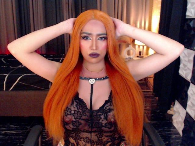 JuicyHugeCockTS with long orange hair, wearing a black lace top, hands in hair, dark makeup, necklace with skull, looking at camera, standing in dimly lit room.