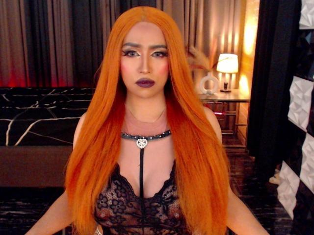 JuicyHugeCockTS with long orange hair, black lace lingerie, and purple lipstick, wearing a black necklace with a silver pendant, sitting in a dimly lit room.