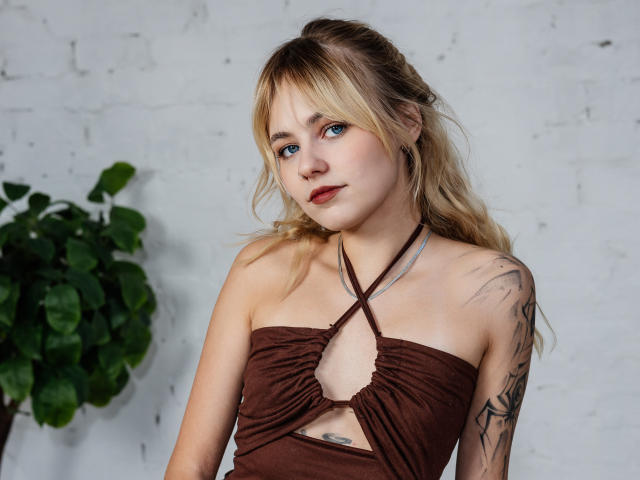 MiaCuteAngel with blonde hair, blue eyes, and tattoos, wearing a brown strapless dress with a cutout, looking slightly to the side, standing next to a green plant.