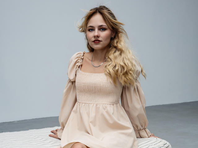 MiaCuteAngel sits on a white, textured surface. She has long, wavy blonde hair, fair skin, and blue eyes. She wears a pale pink, square-neck dress with puffed, sheer sleeves and a cinched waist. Her left hand holds the dress's shoulder strap. The dress has a subtle, textured pattern. She wears a thin, silver necklace. The image is sharp with a neutral, gray background. Her expression is calm, with slightly parted lips.