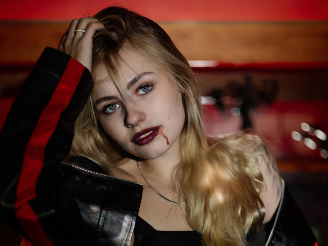 MiaCuteAngel, a blonde woman with blue eyes, wears dark lipstick with a blood-like smear. She's in a black leather jacket with red stripes, her right hand touching her hair. She has a tattoo on her right shoulder. The image is sharply focused on her face, with a wooden and red background blurred. Her expression is intense, and the lighting highlights her facial features.