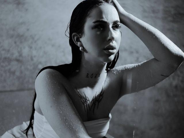 Black and white photo of IsabelleDelicious with wet, dark hair, looking up with a serious expression. She has water droplets on her skin, a tattoo with the word "BOOM" on her chest, and a tree tattoo below her collarbone. She's wearing a strapless top, and her left arm is raised, touching her head. Her right arm is slightly raised, showing a small tattoo on her inner bicep. She has earrings and a subtle makeup look. The photo has a gritty, high-contrast texture.
