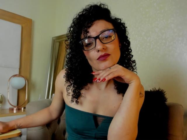 SeleneCollins with curly black hair, glasses, red lipstick, green strapless dress, red nails, hand on chin, looking at camera, tattoo on right wrist, sitting in a cozy room.