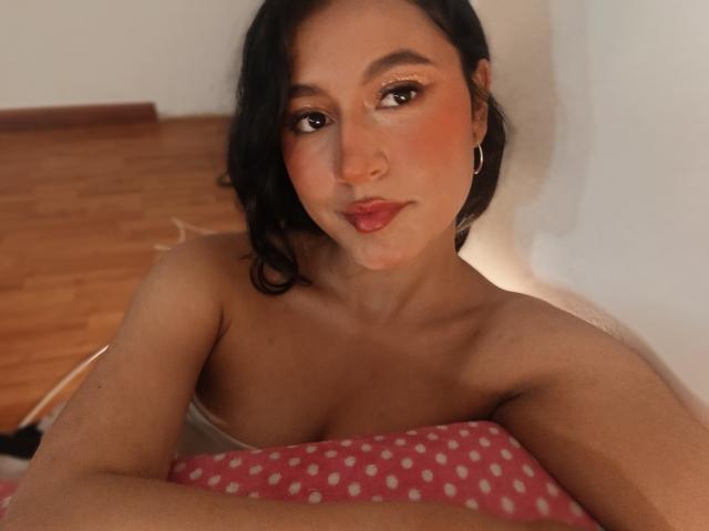 NaughtyAmiyx with medium brown skin, black wavy hair, hoop earrings, pink blush, and red lipstick, topless, holding a pink polka-dot pillow, looking at the camera.