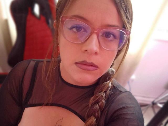 CleareRems with glasses, brown braid, and purple lipstick, wearing a sheer black top, showing a small tattoo on her chest, looking serious. Boobs visible through the top.