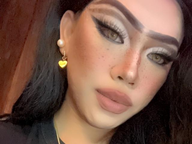 MissCuteBebe has full eyebrows, heavy eyeliner, freckles, and glossy lips. She's wearing a yellow heart earring and a pearl earring, with dark hair and a slightly flushed face.