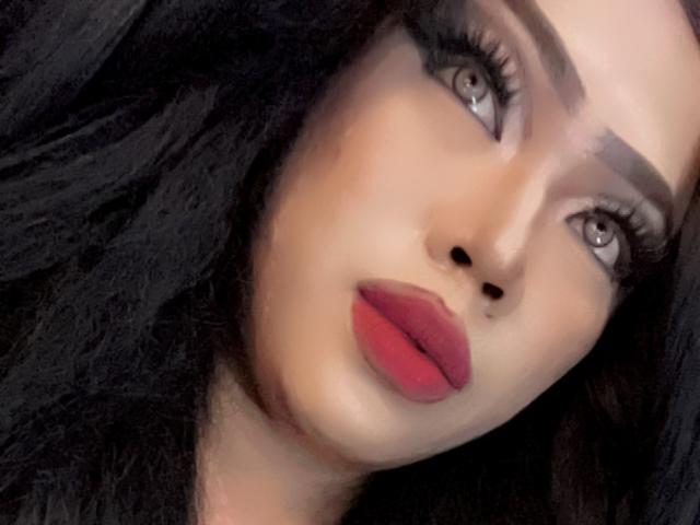 MissCuteBebe's face with dark, wavy hair, gray eyes, thick eyebrows, and red lips. Her makeup is bold, with long eyelashes. Her skin is smooth and light.
