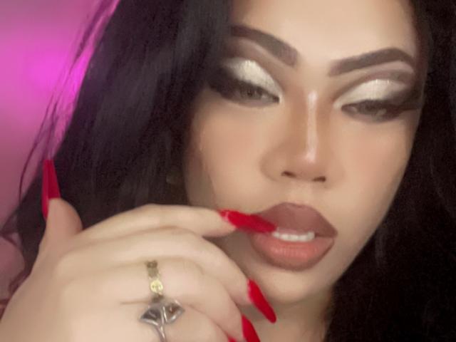 MissCuteBebe's face with glossy silver eye makeup, thick eyebrows, red lipstick, and red nails. She's biting her red nail, wearing a cross ring, with black hair.
