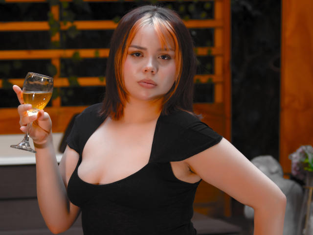 HanniRoss with short brown hair and orange highlights, holding a glass of wine, wearing a black top with a low neckline, showing her tits, looking serious.