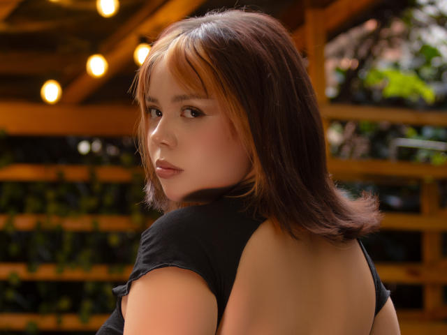 HanniRoss with brown hair and subtle highlights, wearing a black top with a low back, looking over her shoulder with a slightly pouty expression. Her tits are not visible.