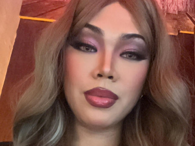 MissCuteBebe has blonde hair, purple eyeshadow, black eyeliner, pink blush, and shiny pink lips. Her makeup is bold and she's looking directly at the camera with a slight smile.