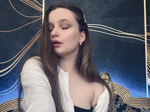 MagikPeachh has pale skin, long brown hair, and closed eyes. She's wearing a white shirt, black top, and showing her tits. Gold lines and dark background.