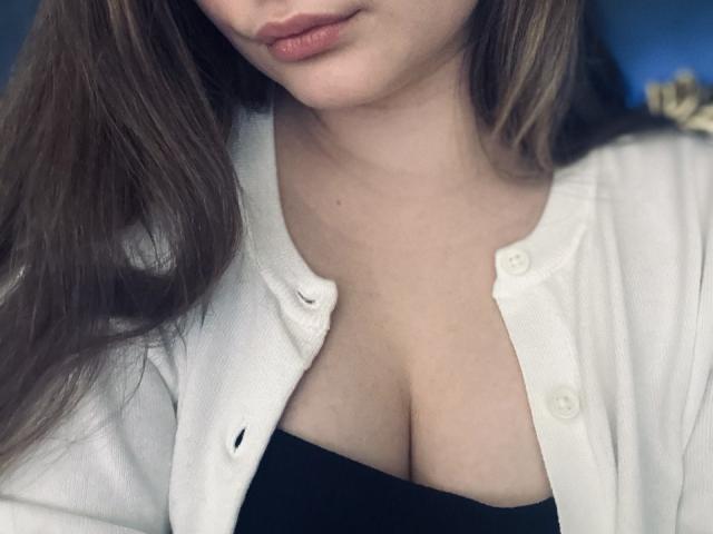 MagikPeachh mit langen braunen Haaren, trägt einen weißen, offenen Cardigan, der ihr schwarzes Oberteil zeigt und ihre Brüste freilegt. Ihre Lippen sind leicht rosa und sie schaut nach unten..