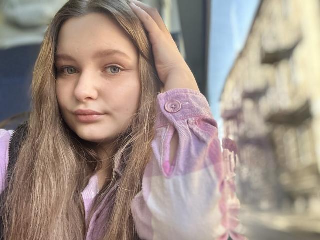 MagikPeachh with long brown hair, pink button-up shirt, and hand in hair, has light green eyes, subtle makeup, and a soft, confident expression. Blurred background.