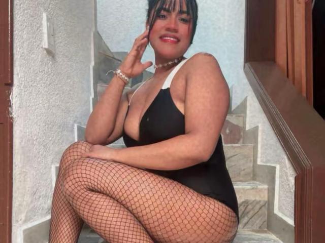 IvannaDuran, with large tits, wearing a black one-piece, fishnet stockings, and a pearl necklace, sitting on stairs, smiling, hand touching her cheek.