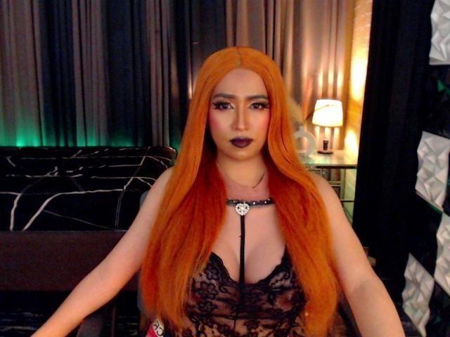 JuicyHugeCockTS with long orange hair, black lace lingerie, dark lipstick, and a black choker necklace, sitting in a dimly lit room with a lamp on a nightstand.