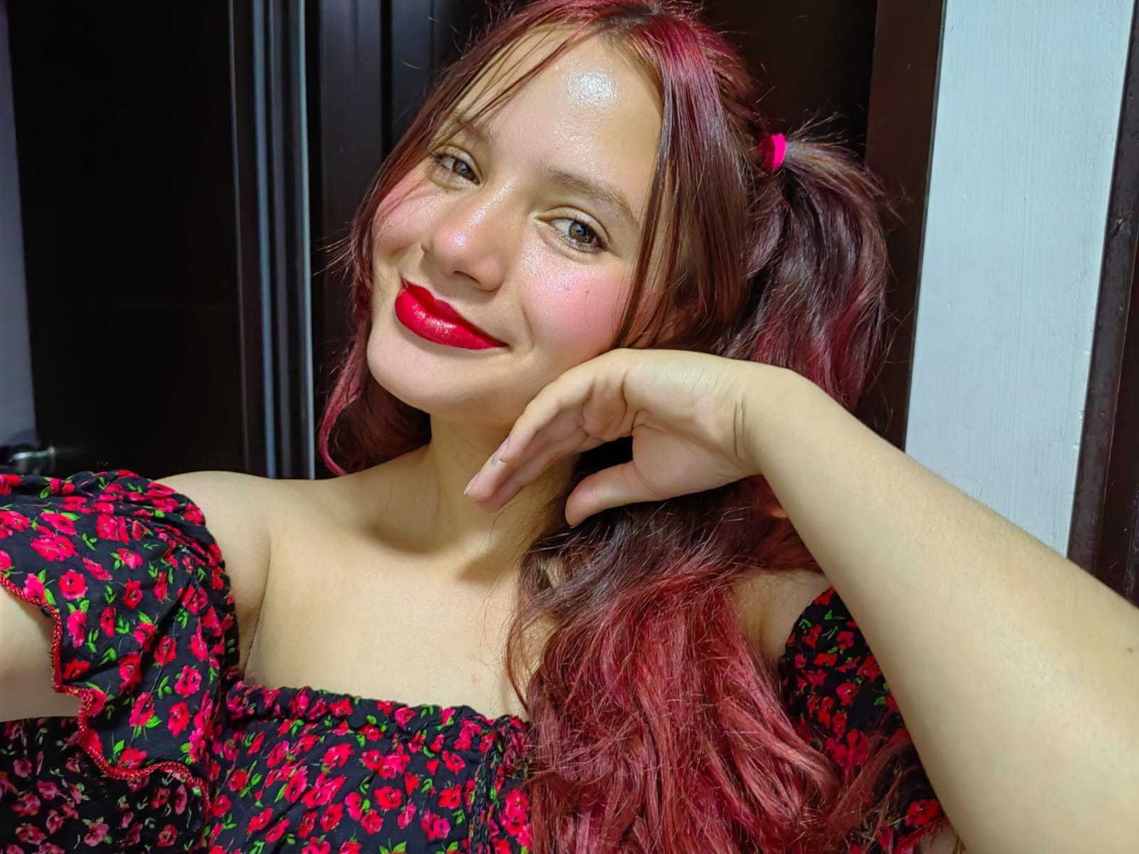 ValeriaWill con lápiz labial rojo, cabello rosa en coletas, vistiendo un vestido floral negro, sonriendo, con la mano en la mejilla, piel clara, rubor rosado, nariz pequeña, busto medio..
