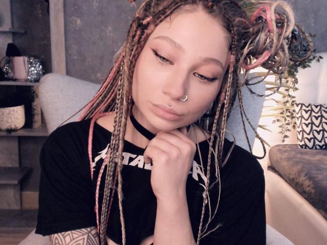 AliceAndCristal has pink and blonde braids, a nose ring, and a black choker. She's wearing a black shirt, looking down with half-closed eyes, and has a tattoo on her arm.