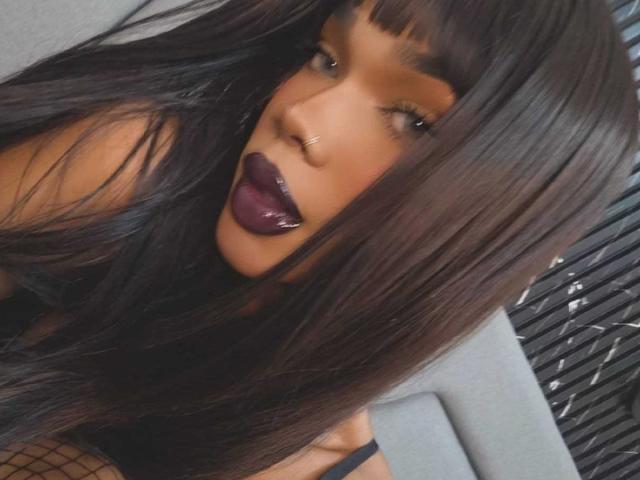 NaomiSteward with long, straight brown hair, dark purple lipstick, and a nose ring, showing her pouty lips and looking up at the camera. Her fishnet top is visible.