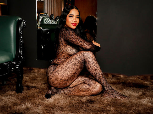KarlaBella with dark hair, red lipstick, and a patterned black fishnet bodysuit, sitting on a furry brown rug, legs curled, arms hugging knees, green chair to the left.
