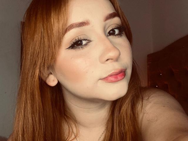 DarkCourtney has pale skin, red hair, and thick eyebrows with black eyeliner. Her pink lips are slightly parted, showing a subtle smile. Her tits are not visible.