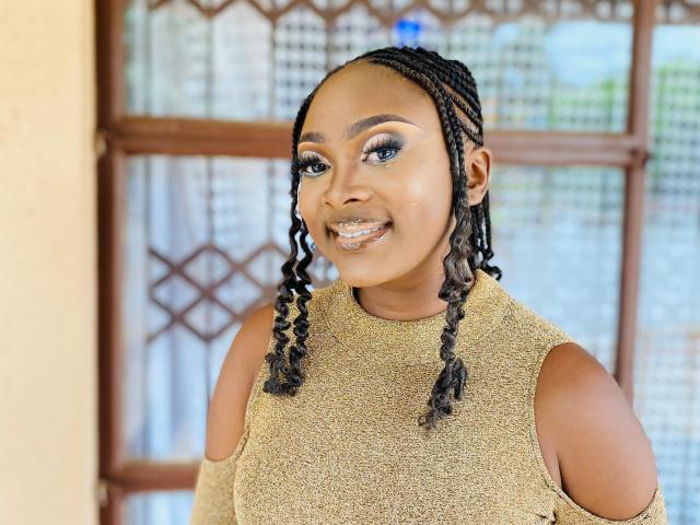 Photograph of StaceyNoelle. She's a dark-skinned woman with braided hair, wearing a textured, beige, sleeveless top with cutouts. Her makeup includes bold, winged eyeliner and shimmering eyeshadow. She's smiling with a silver tooth visible. The background features a wooden window with a geometric pattern and semi-transparent curtains. StaceyNoelle's makeup and hairstyle are detailed and polished, with a confident, approachable expression. The image uses natural light.