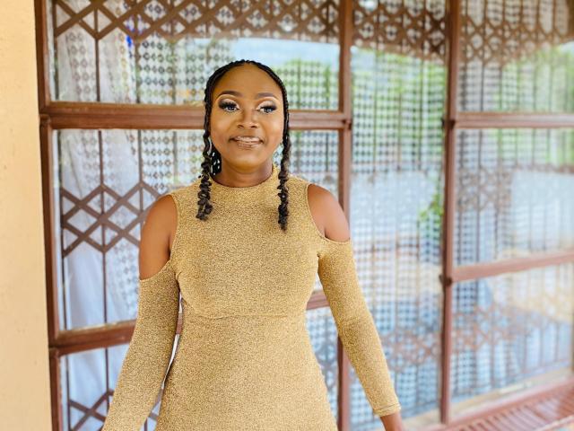 StaceyNoelle stands in front of a wooden lattice, wearing a glittery, tan, off-shoulder dress. She has dark braided hair in two small pigtails, and her makeup includes shimmering eyeshadow and subtle lipstick. Her skin is dark and smooth, and she has a confident, slightly smiling expression. The dress has cut-out shoulders and fits her body snugly, highlighting her curvy figure. The wooden lattice behind her is detailed with geometric patterns, adding texture to the background. The overall composition is balanced and centered, with StaceyNoelle as the focal point.
