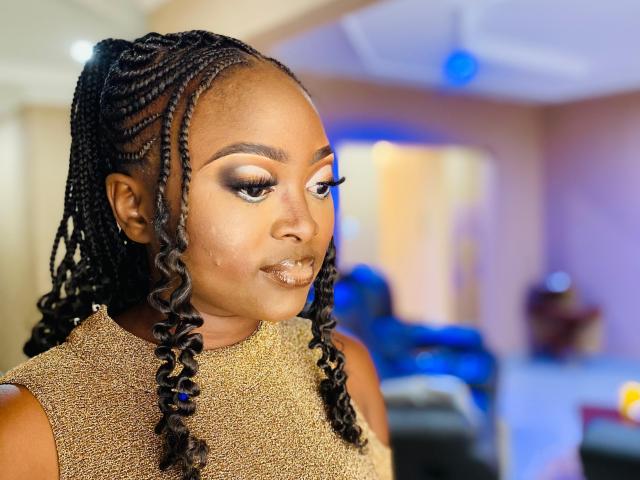 StaceyNoelle, met donkerbruine huid, lang gevlochten haar en opvallende oogmake-up, draagt een gouden jurk met textuur, kijkt naar de zijkant met een lichte glimlach, in een helder verlichte kamer..